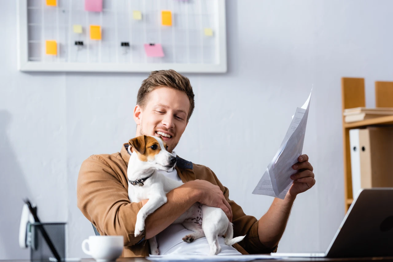 Mann sitzt am Schreibtisch und hält seinen Hund in einem Arm und mit der anderen Hand hält er ein Schriftstück